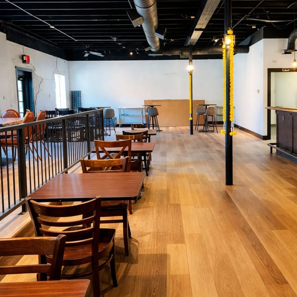 Restaurant dining room with wooden tables and chairs, light wood floor, and black ceiling with exposed ductwork.
