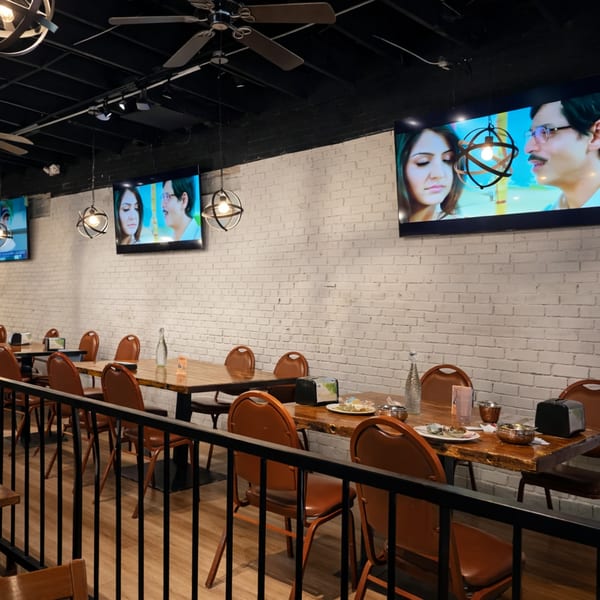 Restaurant dining area with wooden tables, brown chairs, white brick walls, and TVs displaying a movie.